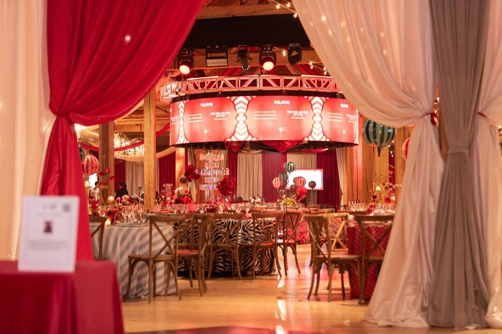 Elegant event space decorated with red and white curtains, a large circular digital display, tables with floral centerpieces, and balloons, set up for a celebration or banquet.