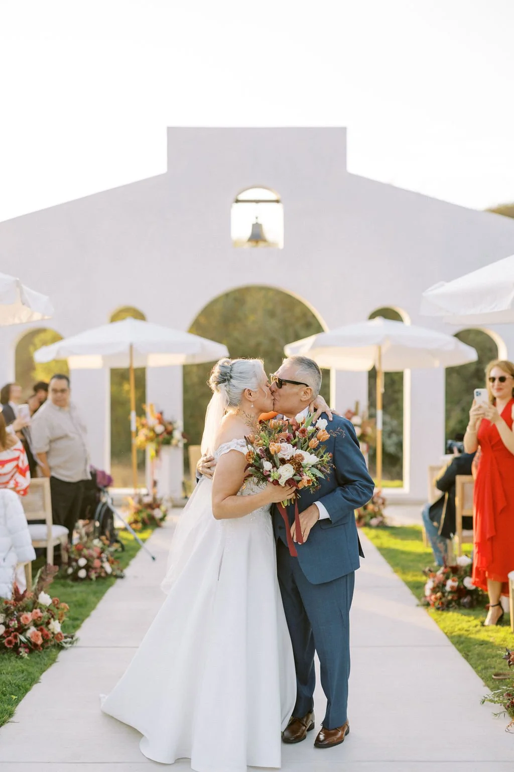 A couple shares a kiss during their outdoor wedding ceremony, with guests and floral arrangements around them.