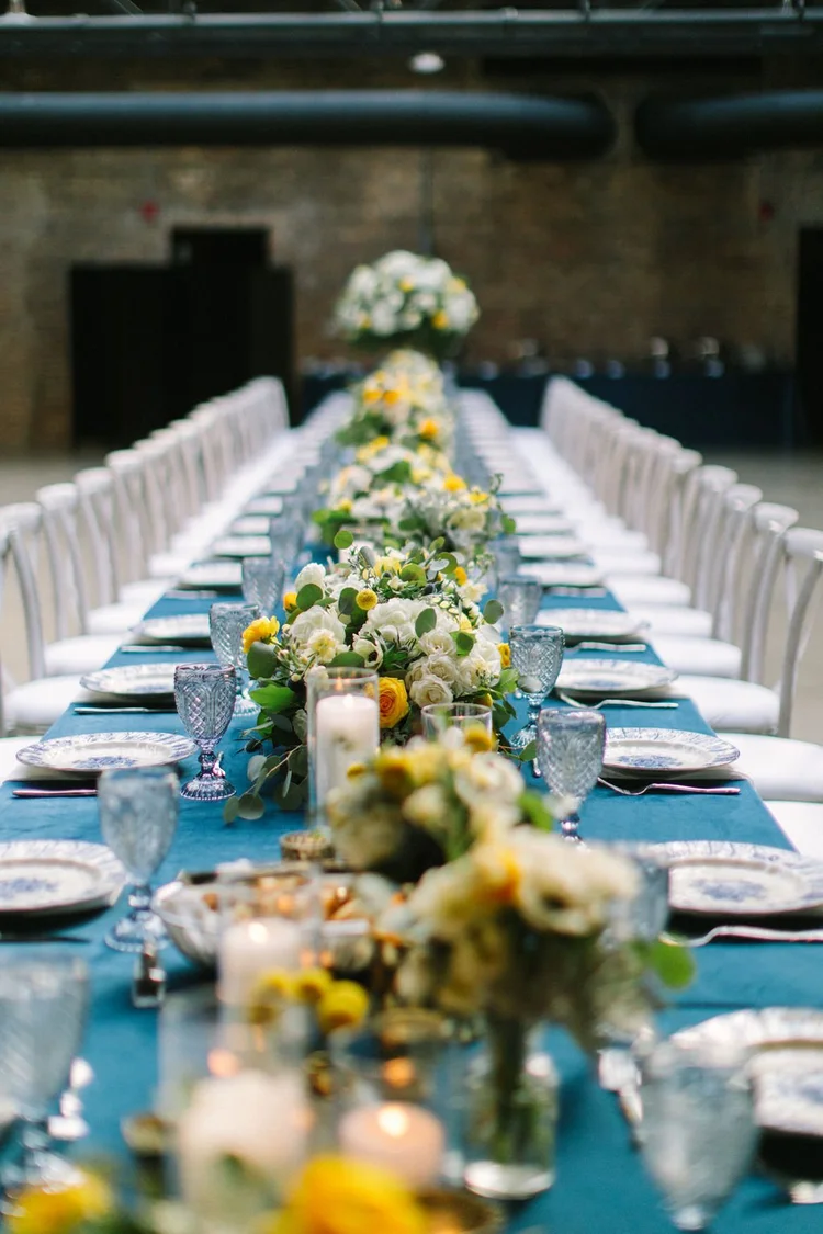 A long banquet table decorated with white and yellow floral arrangements, candles, and blue glassware in an industrial-style venue.