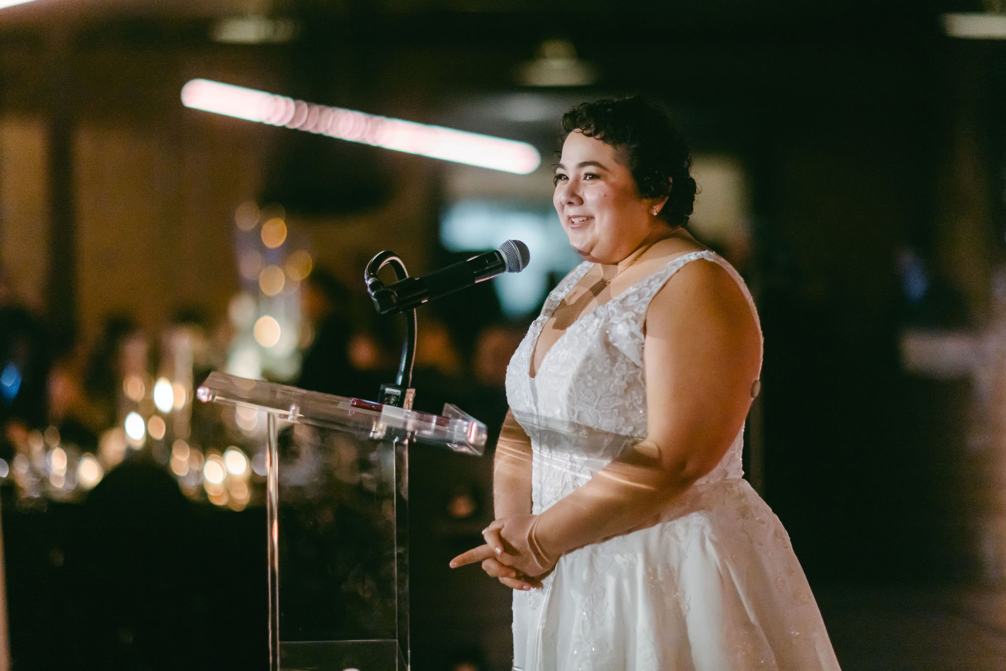 A woman in a white lace dress speaking at a podium with a microphone, smiling at an event with blurred audience and warm lighting.