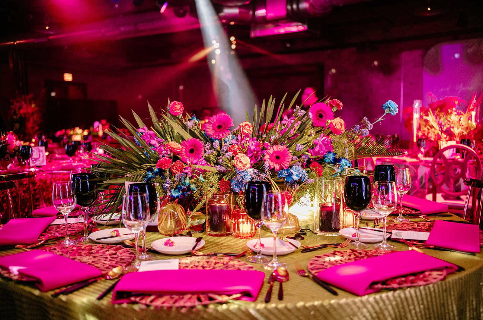 A banquet table decorated with a large pink flower centerpiece, pink napkins, glassware, and candles, set in a dimly lit event space with pink and purple lighting.