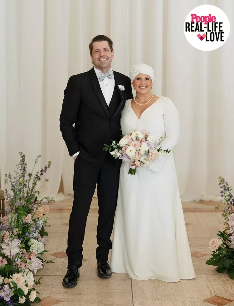 A bride and groom standing together at their wedding, smiling, with the bride holding a bouquet of flowers and the groom in a tuxedo. There are floral arrangements on the floor, and a logo in the top right corner that reads "People Real-Life Love."