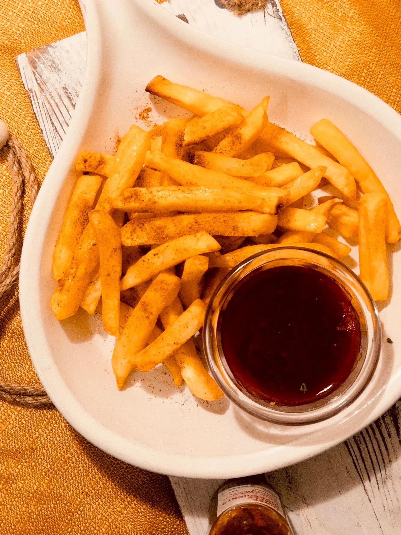 French fries seasoned with paprika served with a small bowl of dark ketchup on a white plate, on a yellow and white striped surface.
