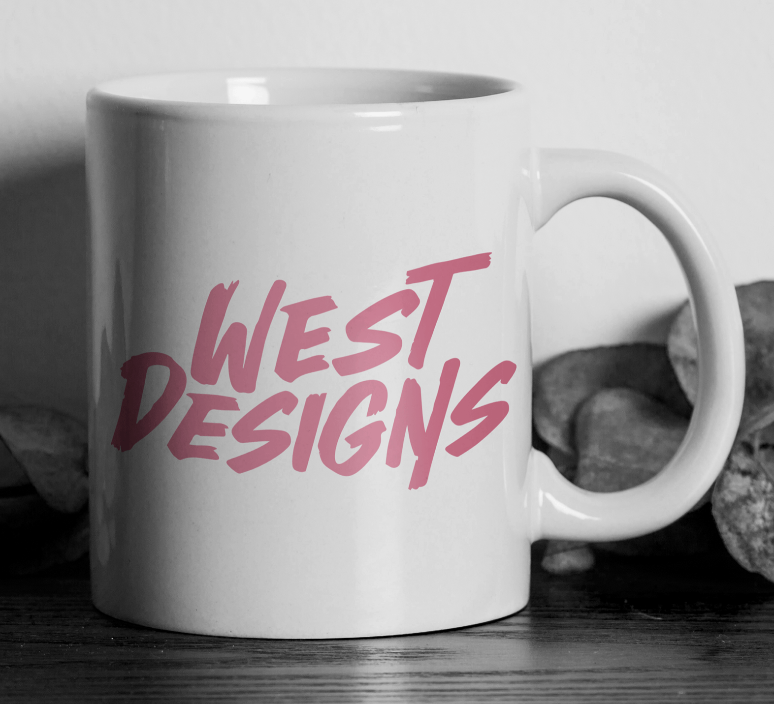 A white ceramic mug with pink text that reads "WEST DESIGNS" on a wooden surface, with rocks in the background.
