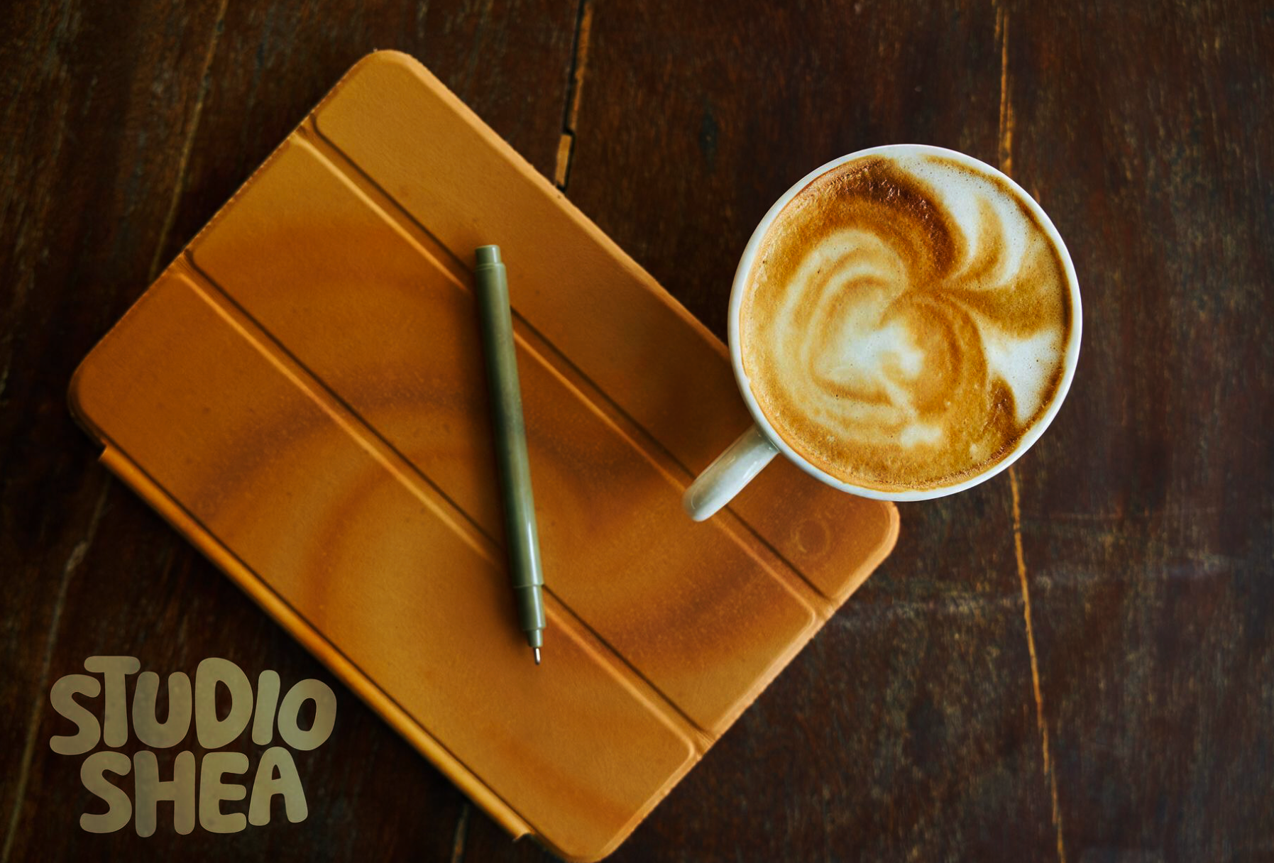 Top view of a coffee mug with frothy latte art, an orange notebook, and a green pen on a dark wooden table.