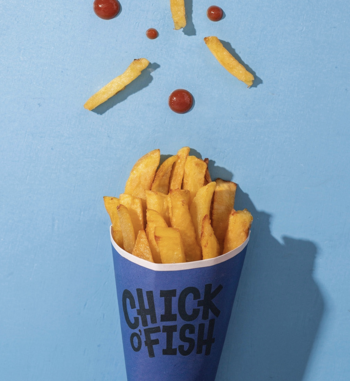 French fries in a blue container with the words "CHICK OF FISH" on it, placed on a light blue surface with ketchup and fry pieces arranged in a face-like pattern above the container.