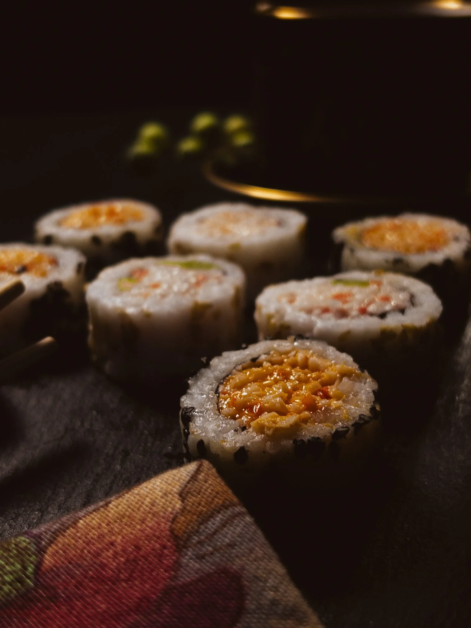 Close-up of sushi rolls with rice, vegetables, and possibly seafood, placed on a dark surface with a colorful cloth underneath.