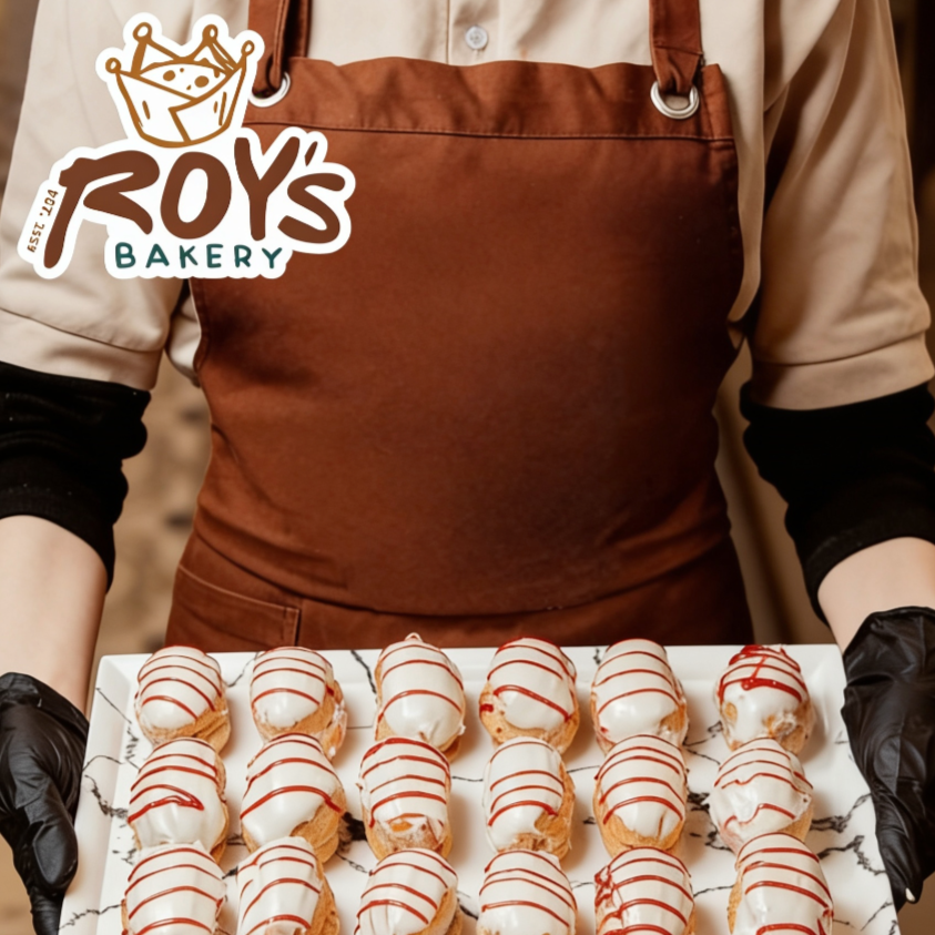 Bakery employee holding a tray of decorated cream-filled pastries topped with white icing and red drizzle at Roy's Bakery