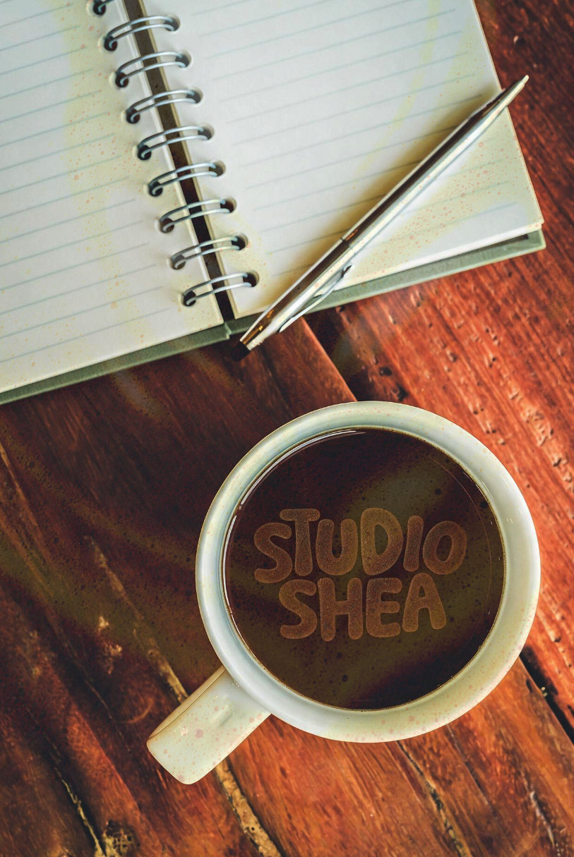 An open spiral notebook with lined pages, a silver pen resting on top, and a cup of coffee with the words 'STUDIO SHEA' written in the foam, all on a wooden table.
