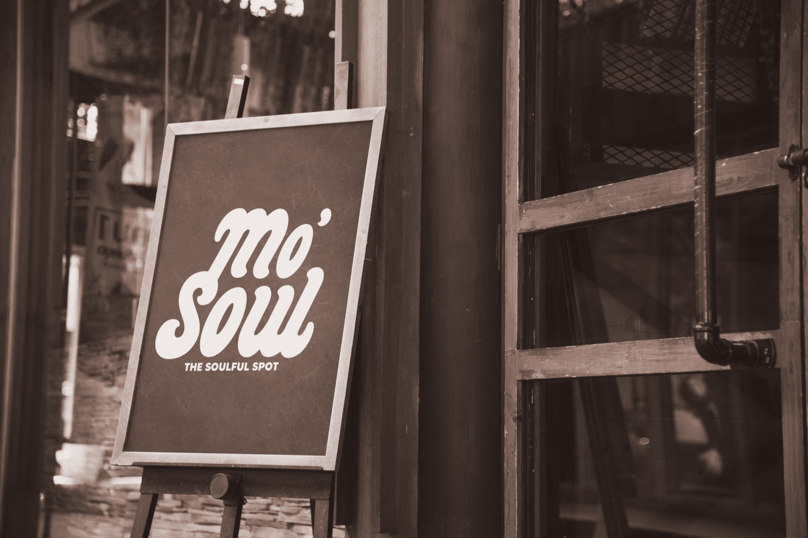 A chalkboard sign outside a shop reads 'mo' soul' and 'the soulful spot' in white text. The shop has a wooden exterior with a glass window and a black pipe is visible. The image is in sepia tone.