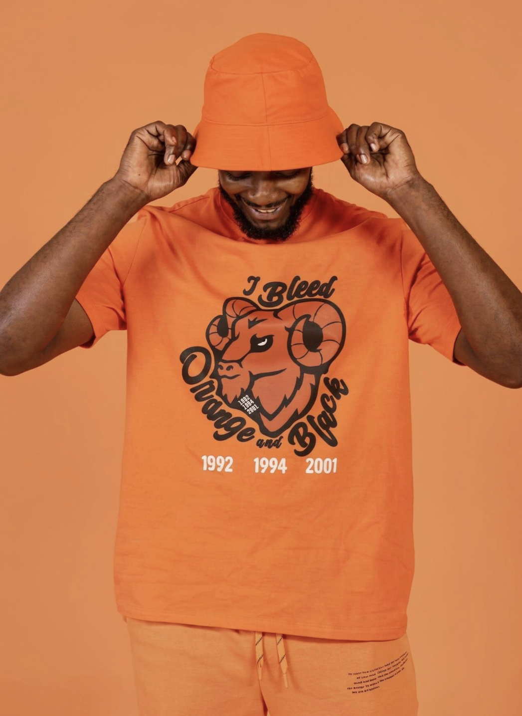 Man in orange clothing adjusting orange bucket hat, wearing T-shirt with a goat illustration and the text 'I Bleed Overtime and Blood' along with the years 1992, 1994, and 2001.