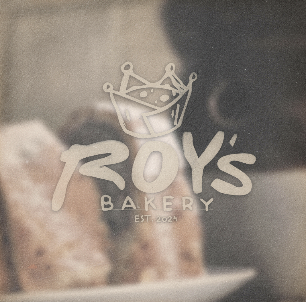 Logo for Toy's Bakery with a crown above the text, established in 2024, on a glass surface reflecting baked goods in the background.