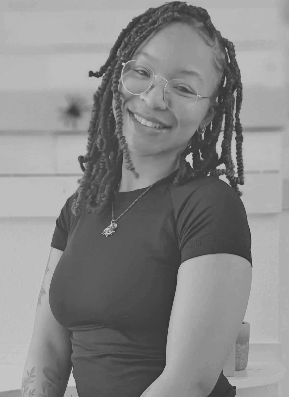 A smiling woman with glasses and dreadlocks, wearing a black shirt and necklace, posing in front of a horizontal wooden wall.