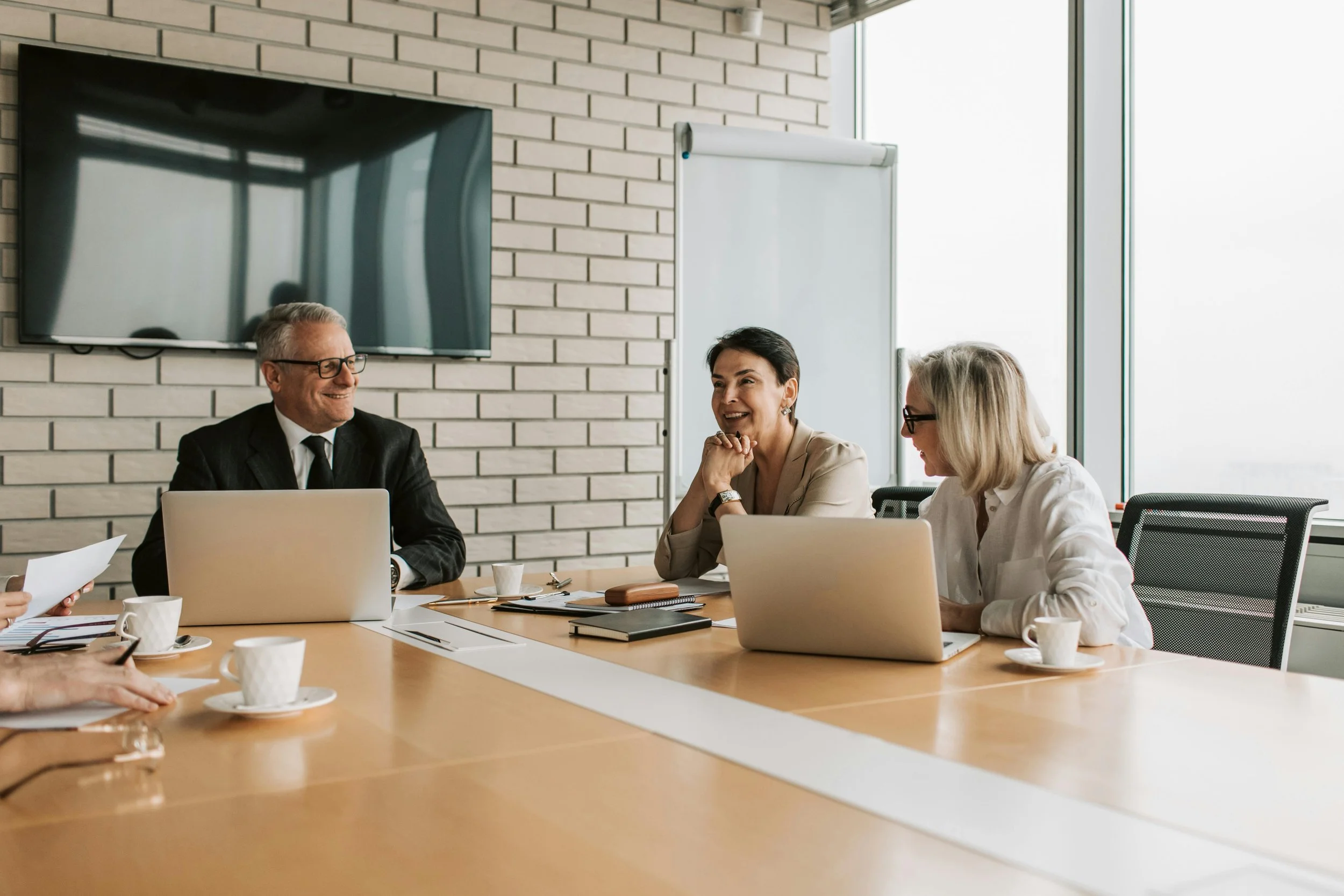 Business meeting with four people sitting around a conference table, two women and one man in a suit visible, with notebooks, laptops, and coffee cups, in a bright office with large windows and a whiteboard.