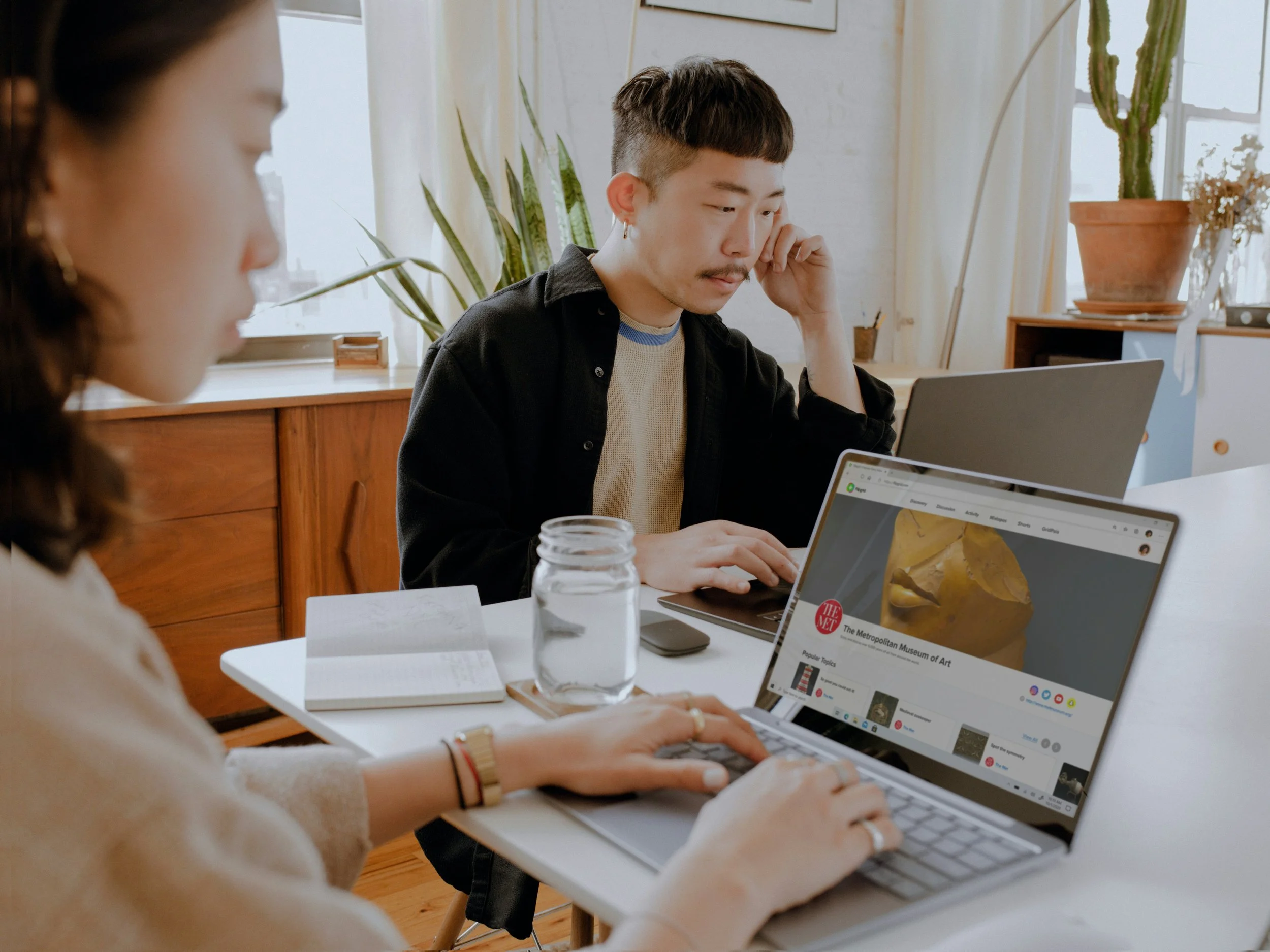 Two people working on laptops in a bright room with plants and books on a table.