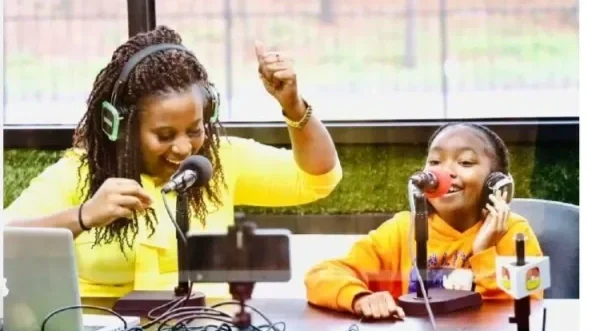 A young woman with long dark hair, wearing a white long-sleeve shirt and yellow pants, standing in front of a glass door entrance, holding a silver laptop, smiling, with blurred people walking behind her.