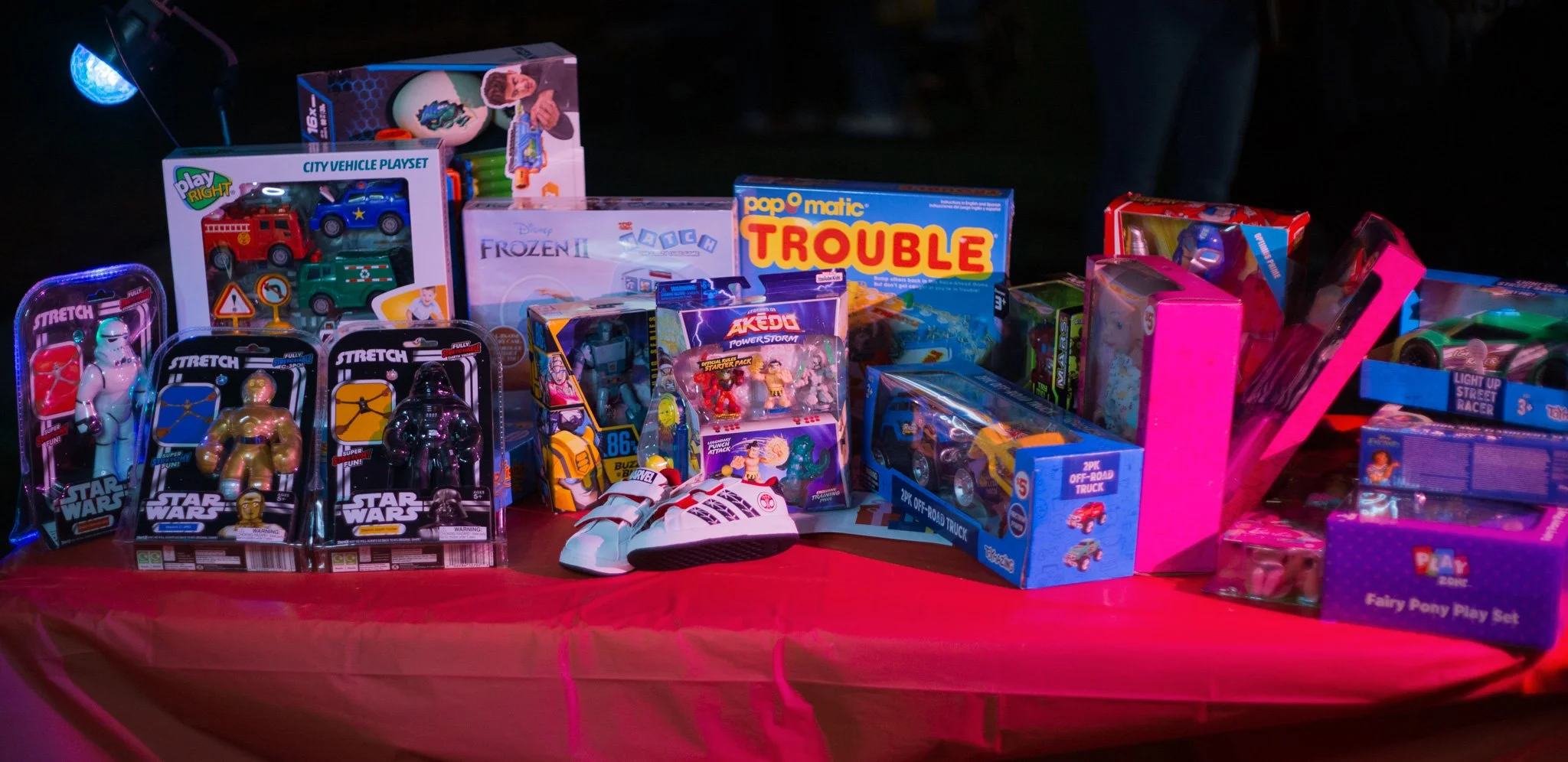 Table filled with children's toys including Star Wars action figures, vehicle playsets, Disney Frozen II toy, board games like Trouble, remote control trucks, dolls, and a fairy pony play set, illuminated by colorful lights.