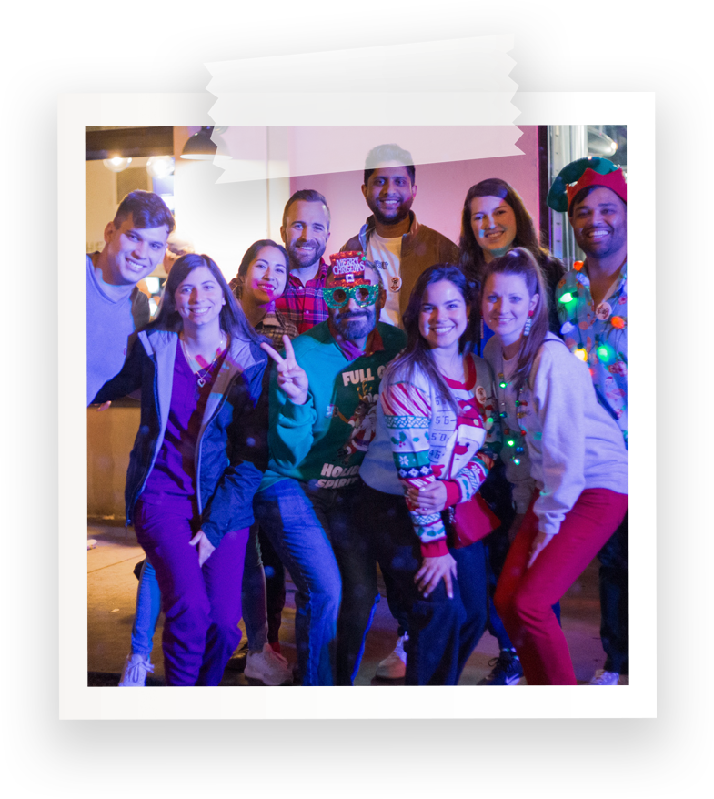 Group of nine people celebrating with Christmas themed and festive attire, some wearing holiday sweaters and accessories, indoors with warm lighting.