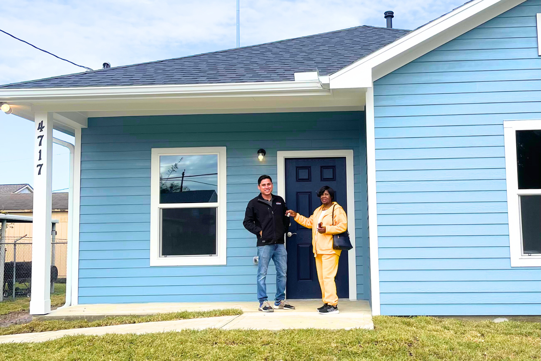 A man and woman standing in front of a blue house. The man is holding keys and the woman is holding a phone. The house has blue siding, a dark blue door, and two white-framed windows.
