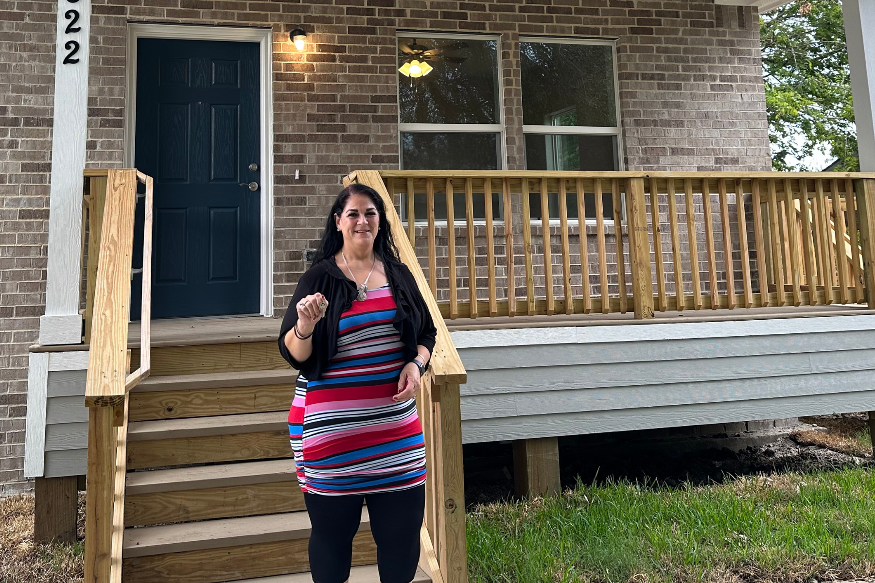 A woman standing in front of a house with a newly built wooden porch and stairs. The house has a brick exterior with a black door, two windows, and a porch light turned on.