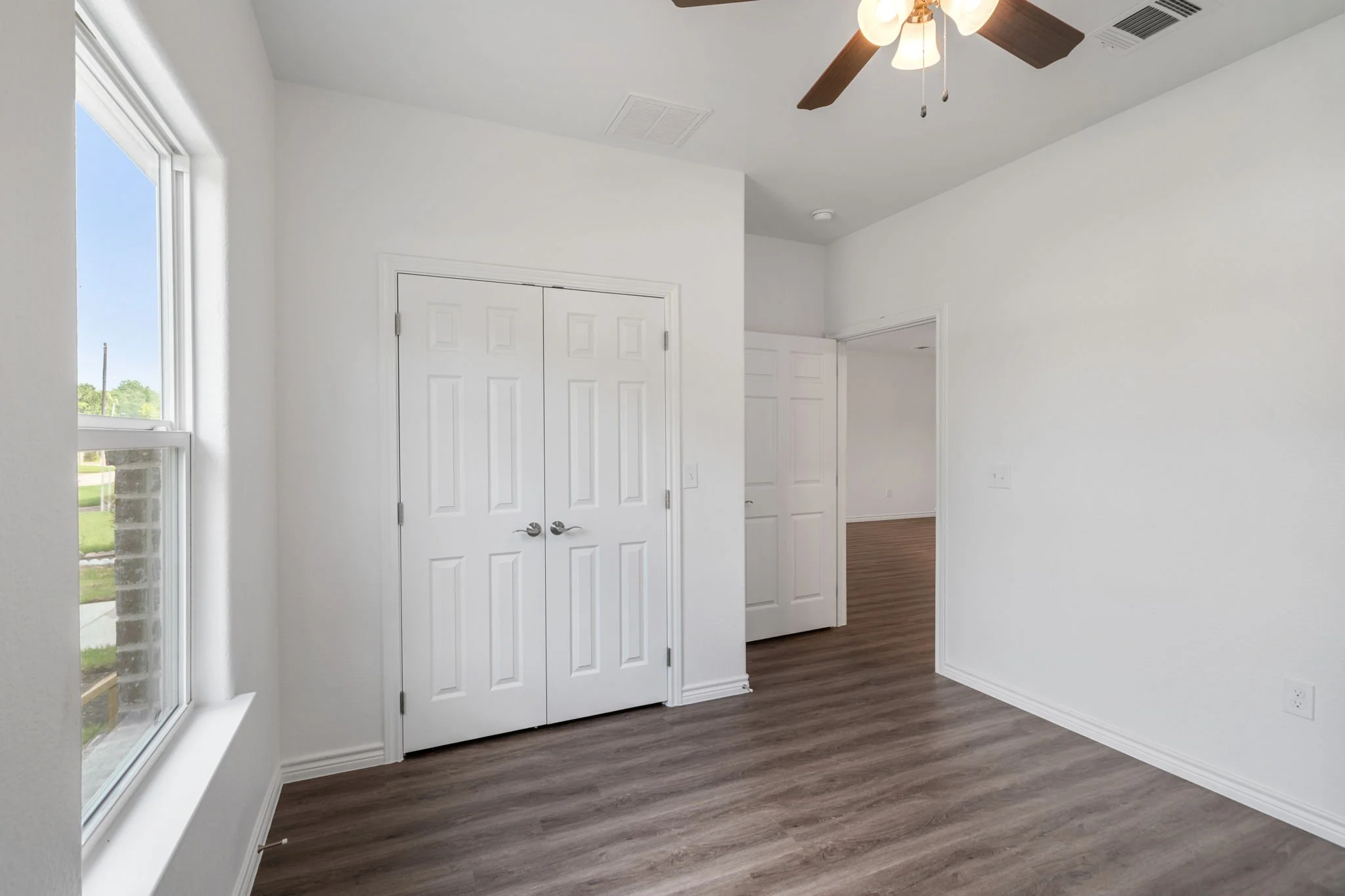 Empty room with white walls, hardwood flooring, a double closet door, a window, and a ceiling fan.
