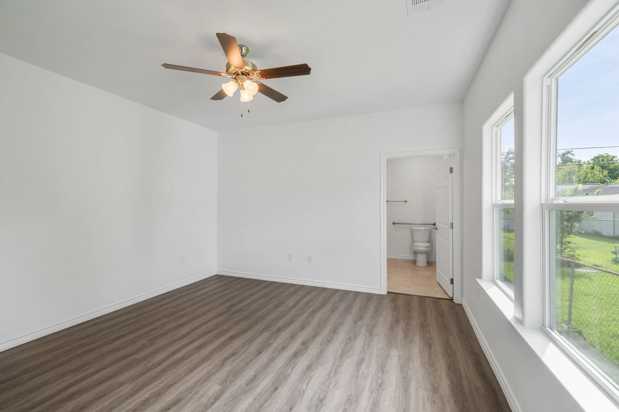 Empty bedroom with wood flooring, white walls, large windows, ceiling fan with lights, and an open bathroom door showing a toilet.