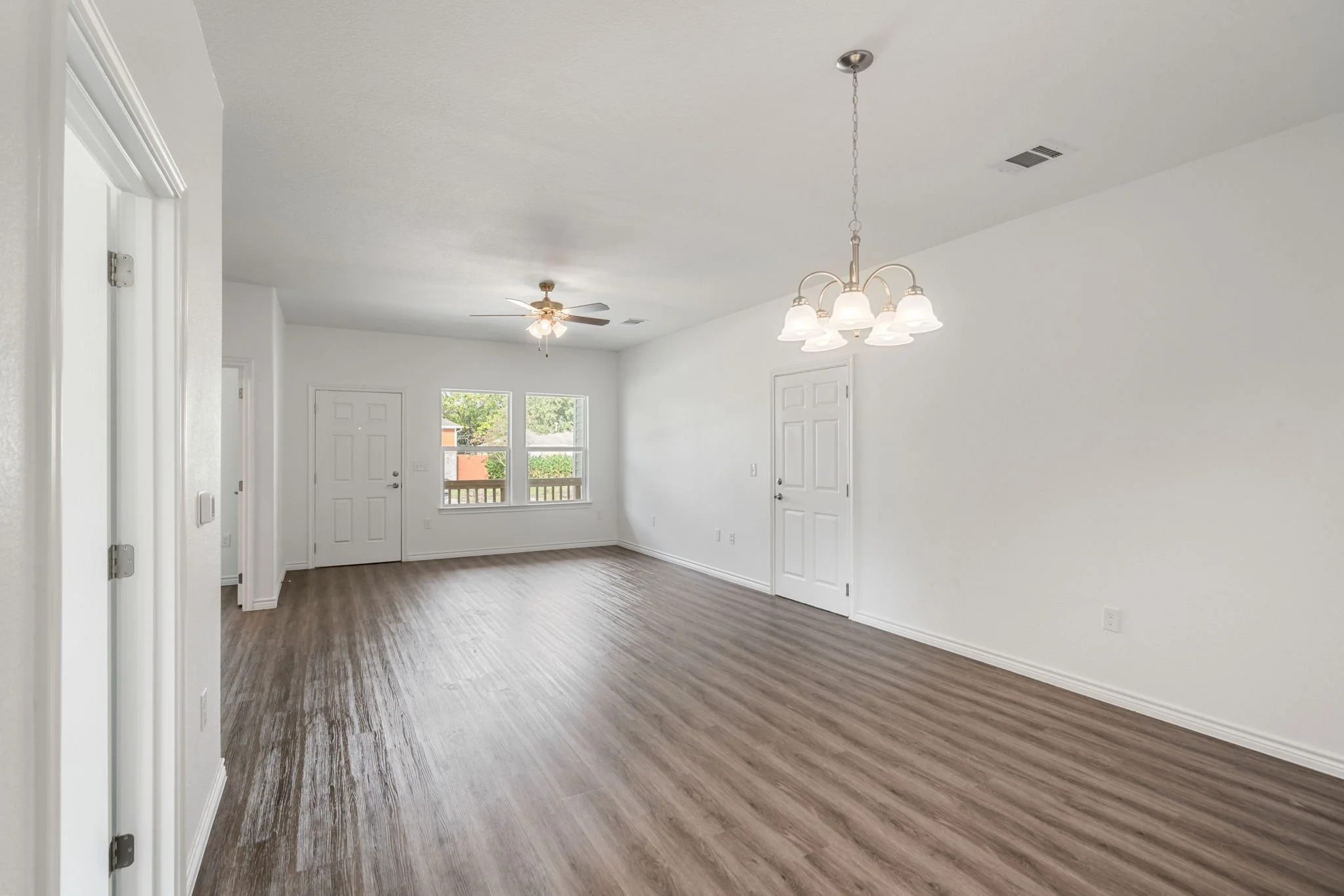 Empty living room with white walls, wood flooring, ceiling fan, and a chandelier, with windows and a door to outdoors.