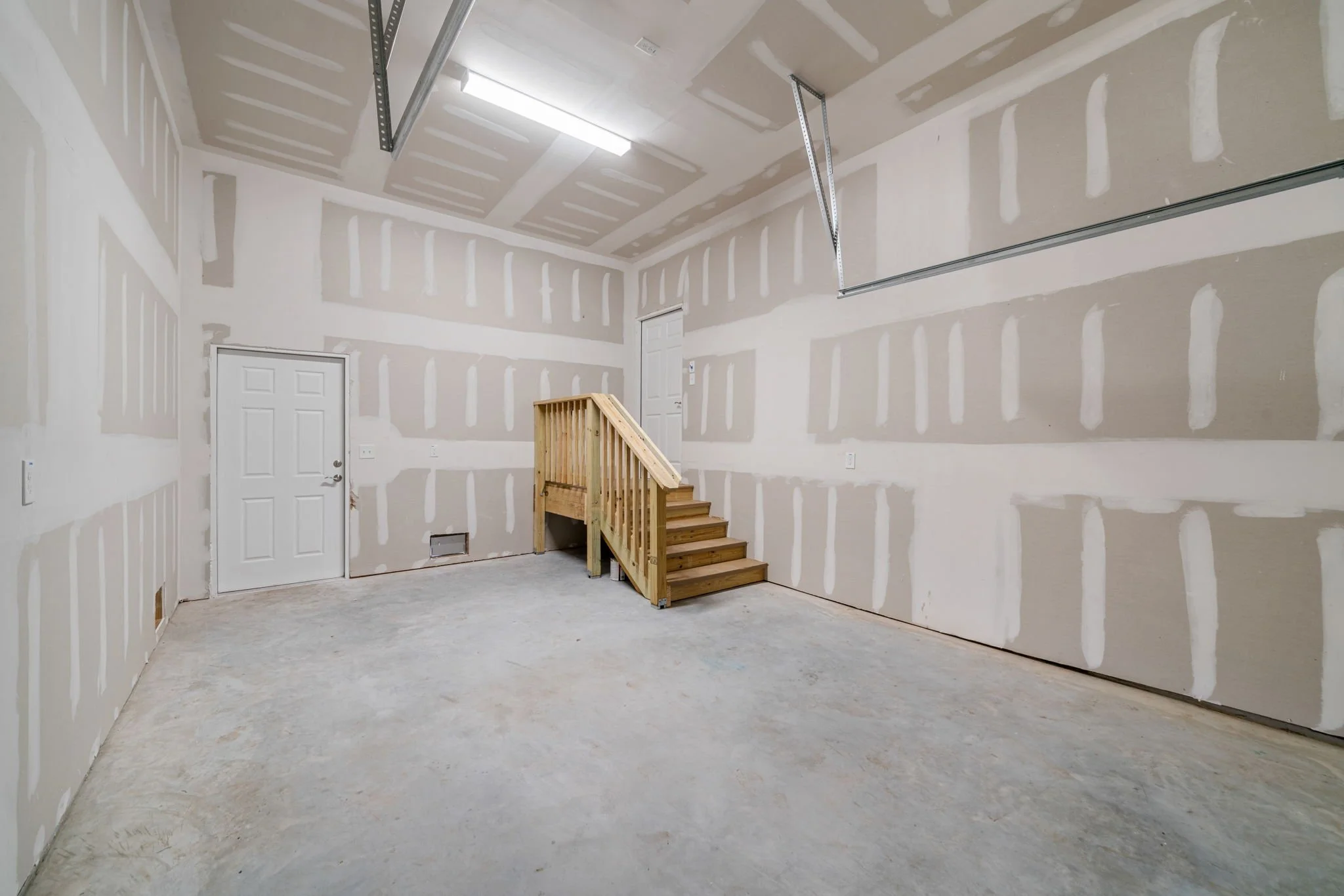 Empty garage with unfinished drywall walls, ceiling, and concrete floor. There is a small wooden staircase leading to a door inside the garage. The ceiling has a fluorescent light fixture and a garage door opener rail.