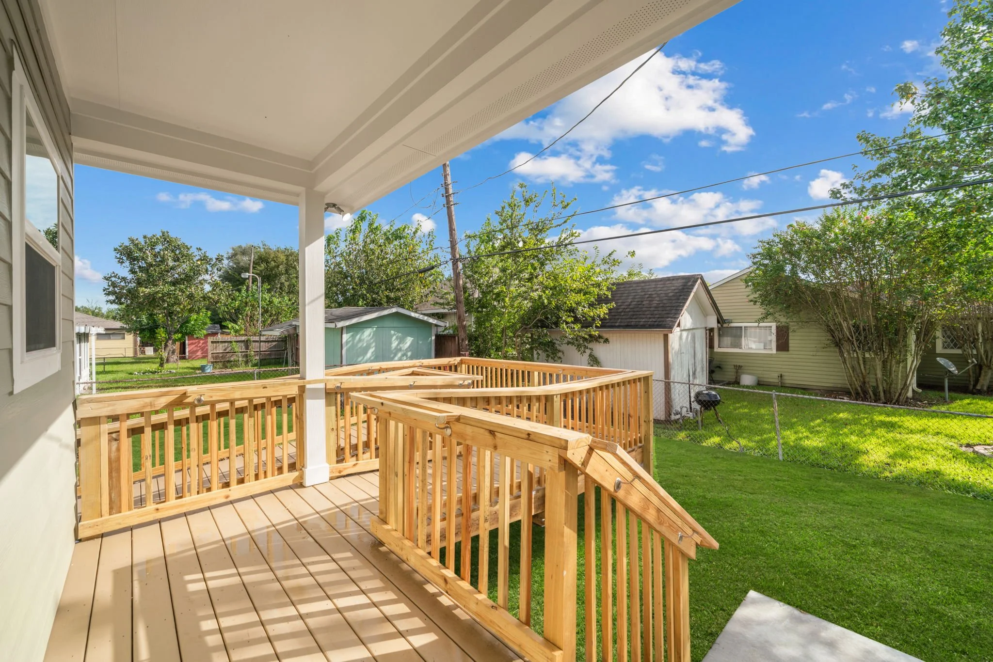 View from a house porch showing a newly built wooden deck, green lawn, trees, neighboring houses, and a partly cloudy sky.