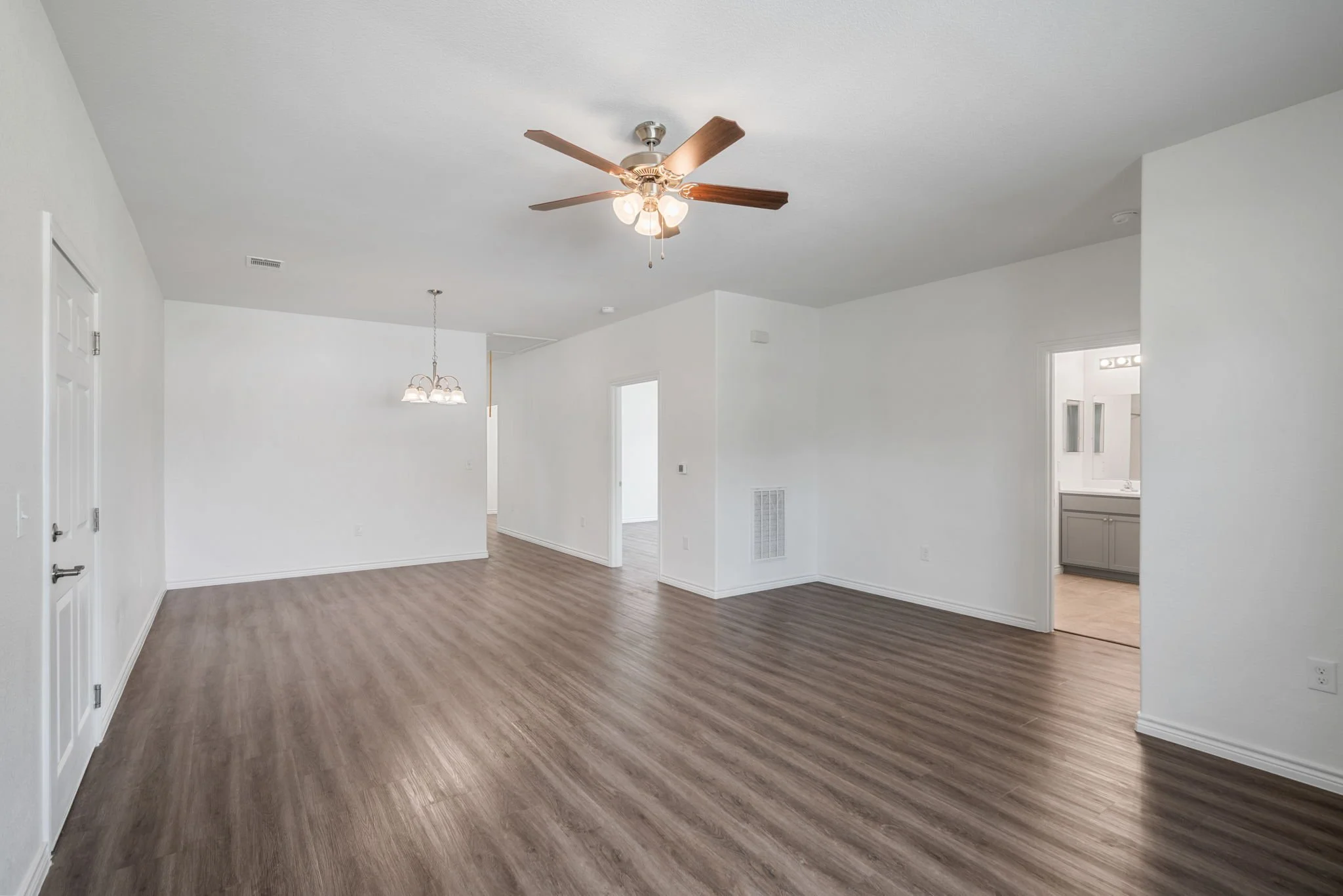 Empty living room with white walls, wood laminate flooring, ceiling fan, and a chandelier over the dining area, with doorway leading to a bathroom.