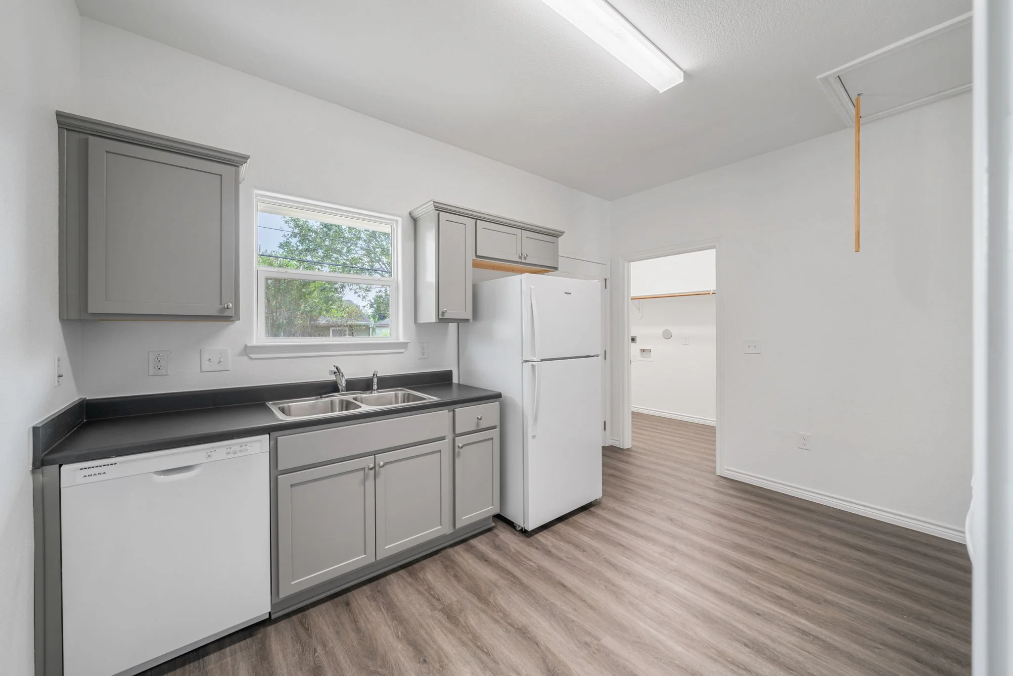 Clean kitchen with gray cabinets, black countertop, white appliances, hardwood floor, and a window over the sink.