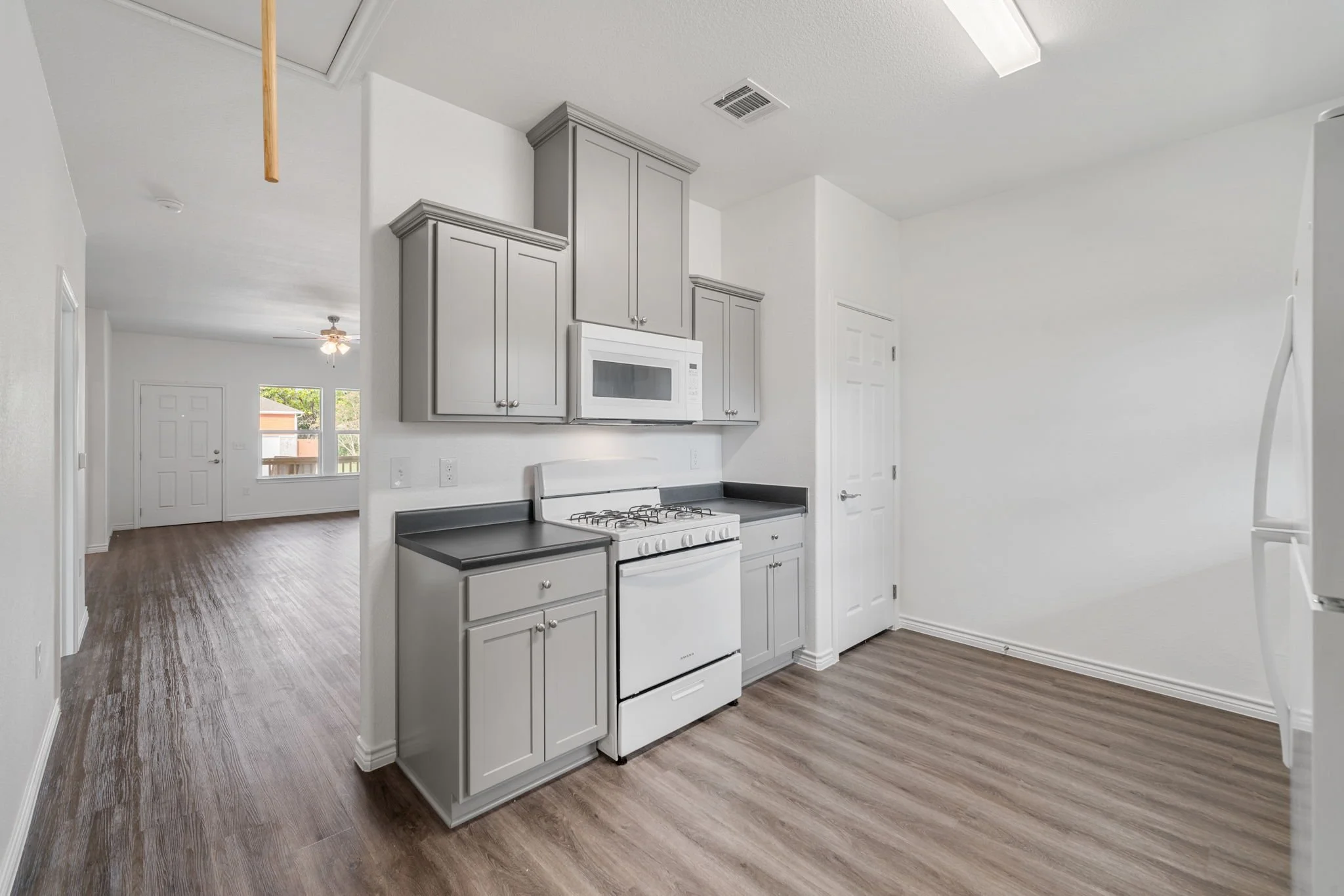 Empty kitchen with gray cabinets, white appliances, and wood flooring, with a view into a living room with a ceiling fan and a window.
