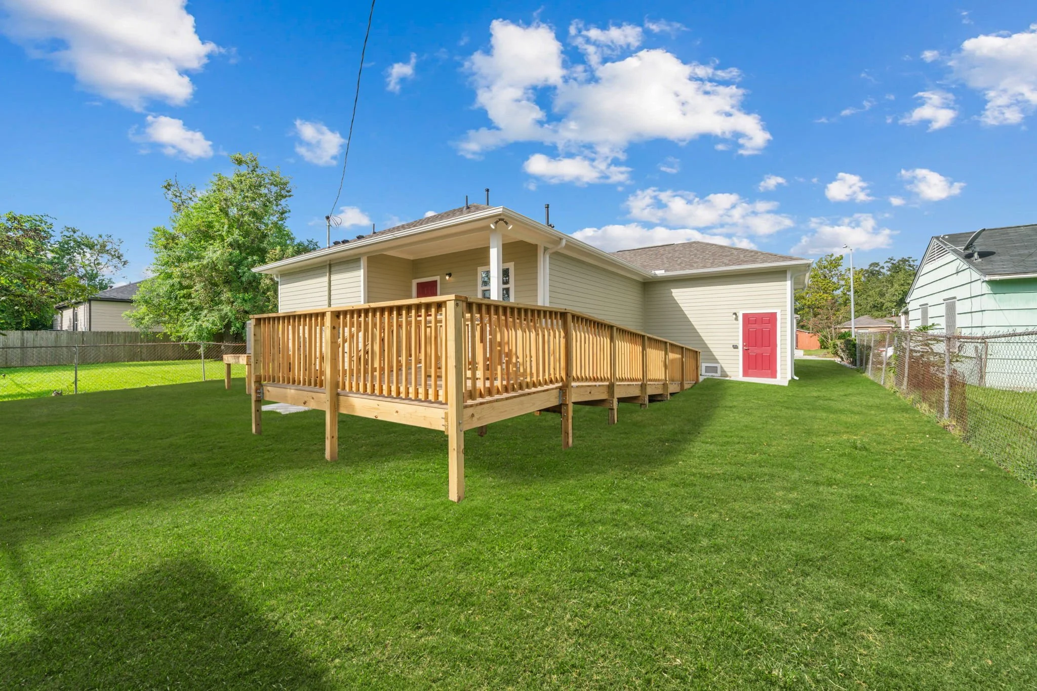 Backyard view of a house with a newly constructed wooden deck and green grass, under a blue sky with scattered clouds. The house has beige siding and red doors.