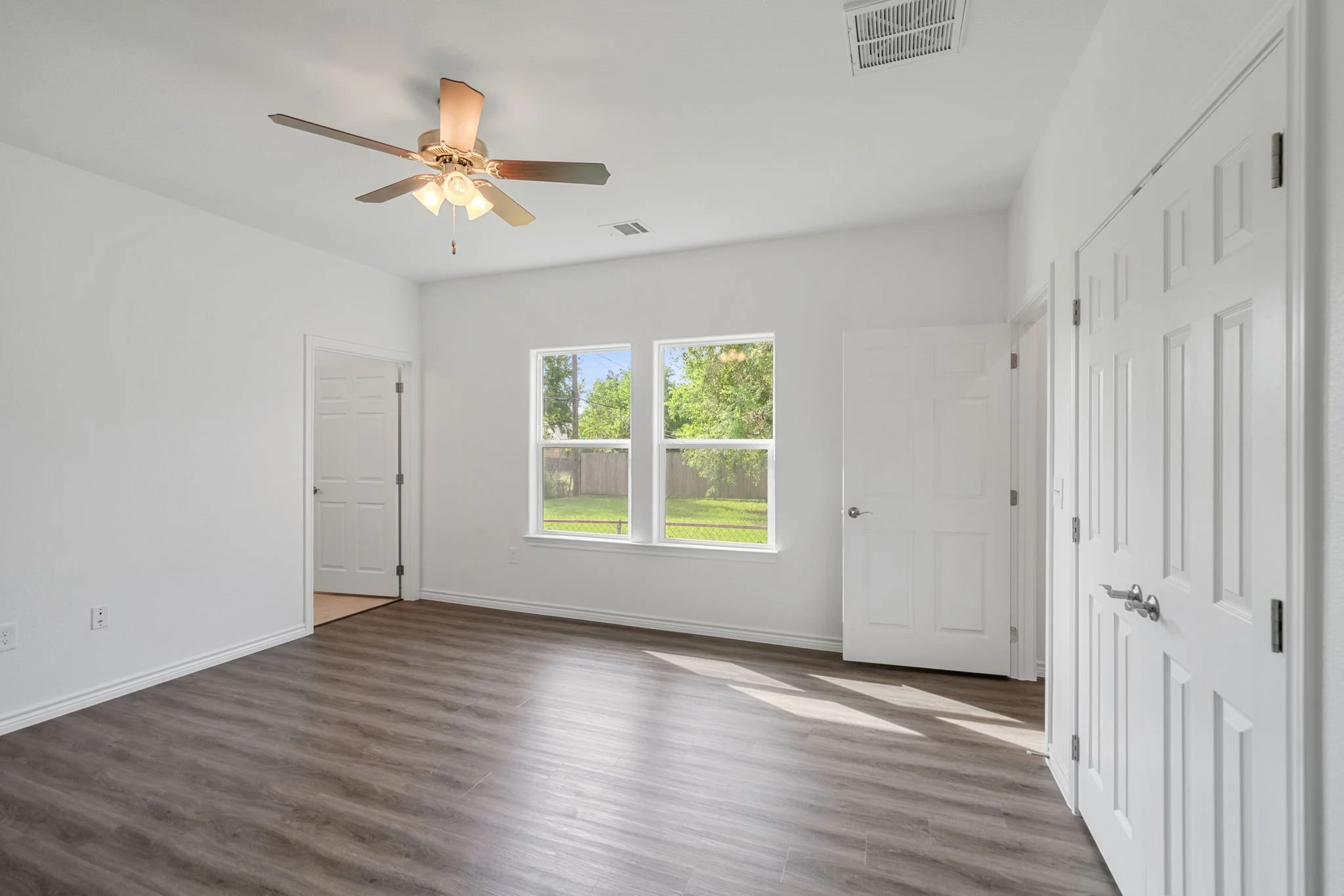 Empty room with white walls, wood flooring, two windows showing a green yard, a ceiling fan with lights, multiple doors, and ceiling vents.