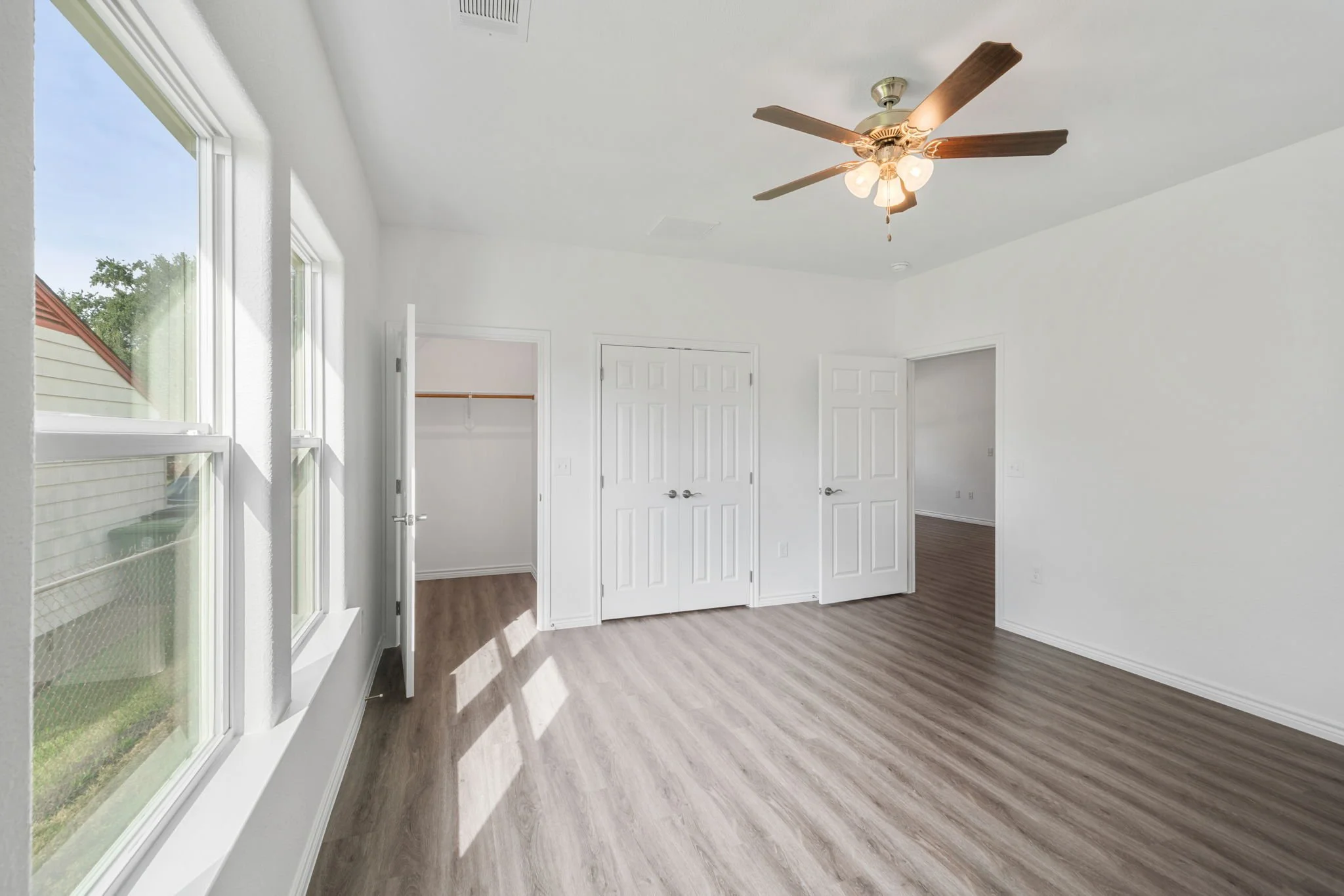Empty bedroom with white walls, wood vinyl flooring, a ceiling fan with lights, three windows, and two open closet doors.