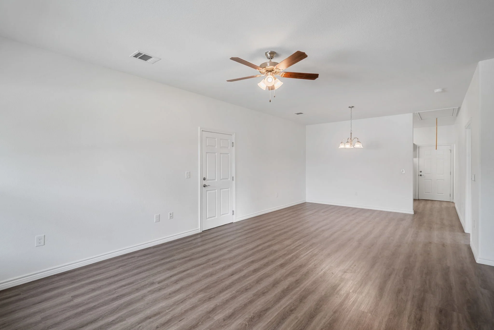 Empty living room with hardwood floors, white walls, ceiling fan, and a hanging light fixture near the entrance.