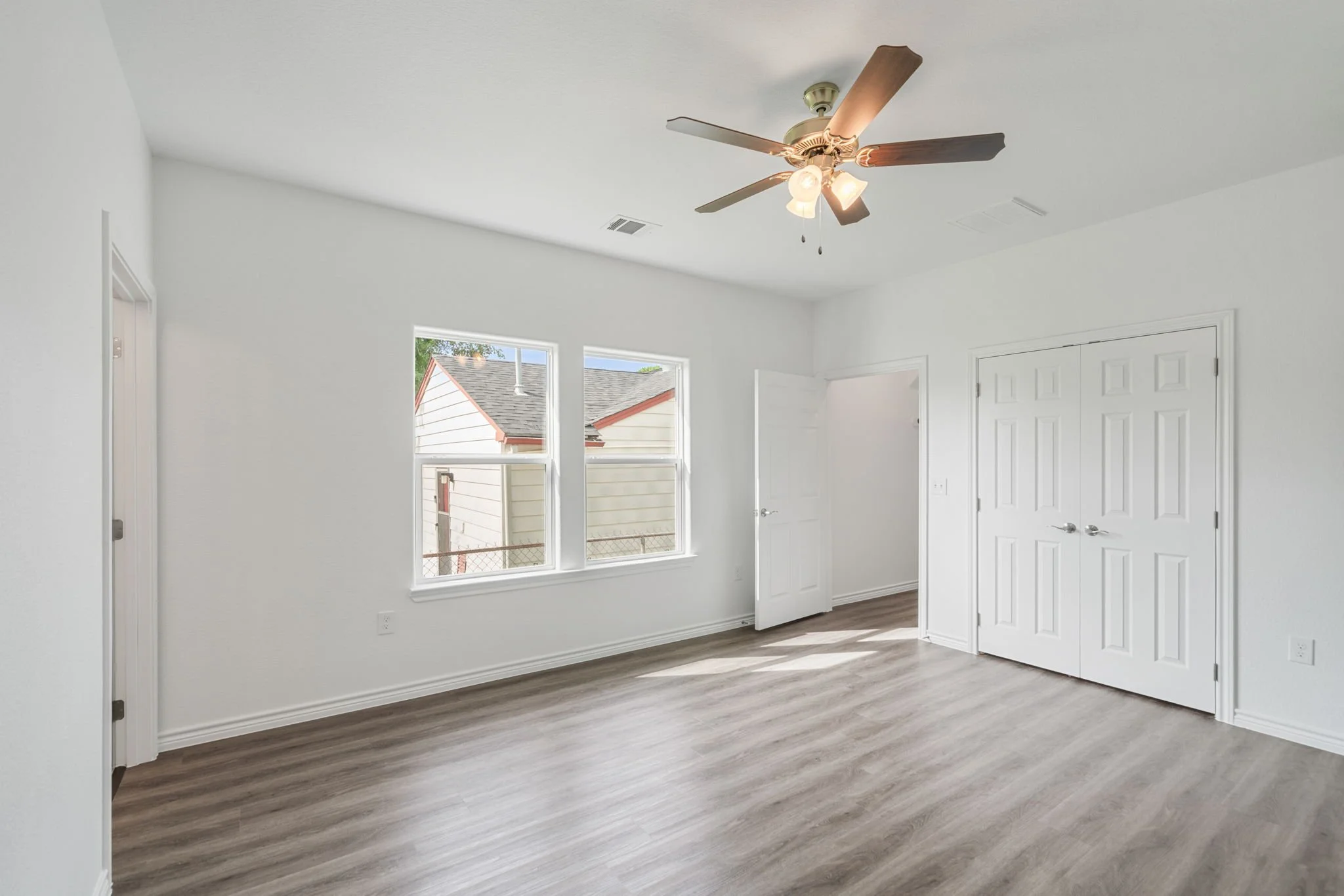 Empty bedroom with white walls, wooden flooring, a ceiling fan, two windows, and a closet with double doors.