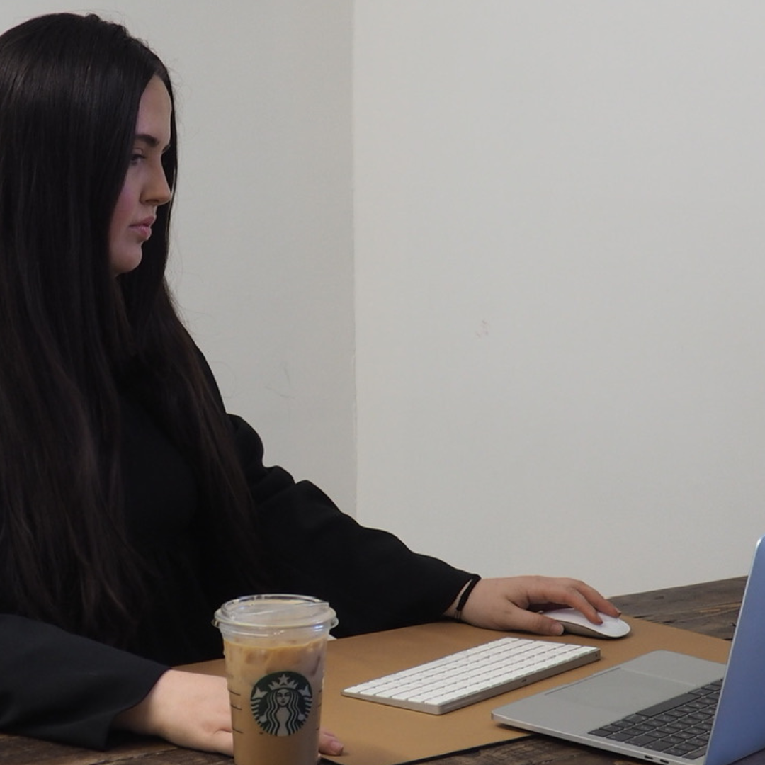 A young woman with long dark hair working on a laptop at a wooden table, with a Starbucks iced coffee nearby.