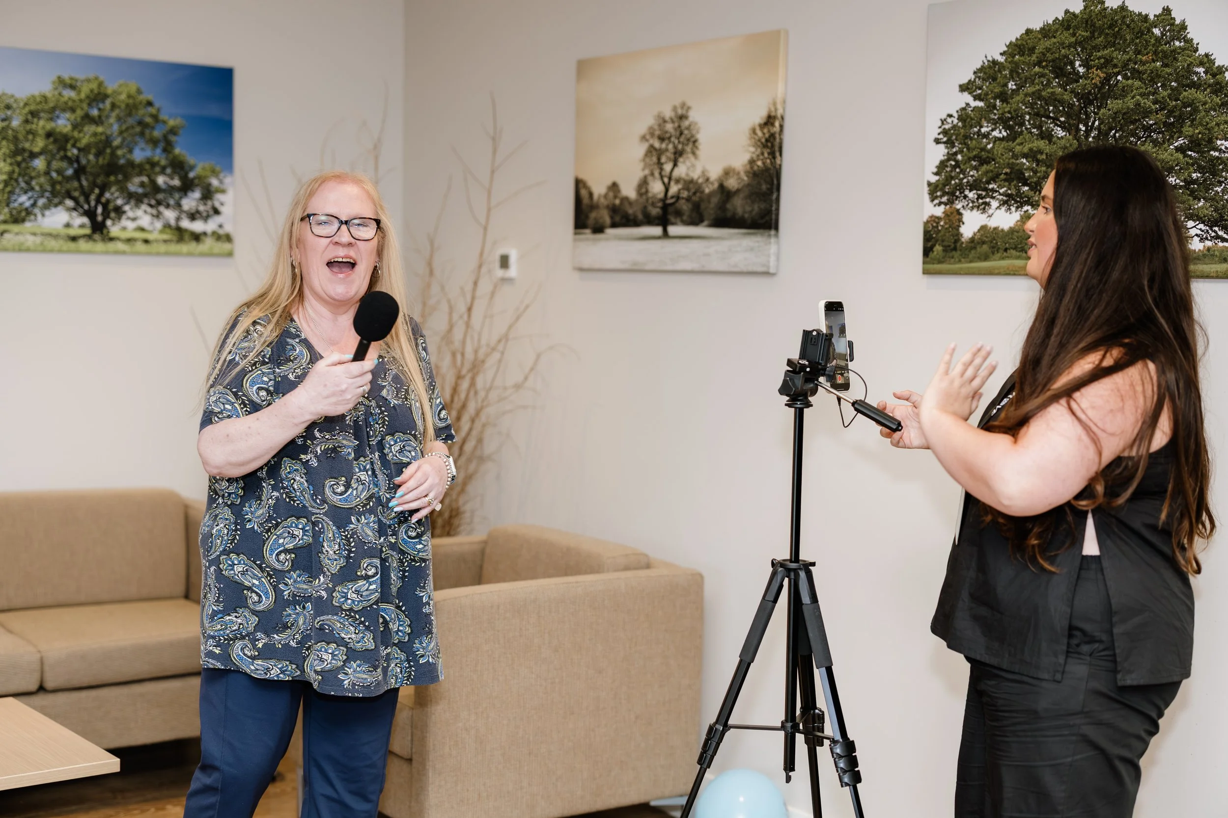 A woman with long blond hair and glasses singing into a microphone, with a woman with long dark hair and a black outfit recording her with a smartphone on a tripod, in a room with beige walls and framed photos of trees.