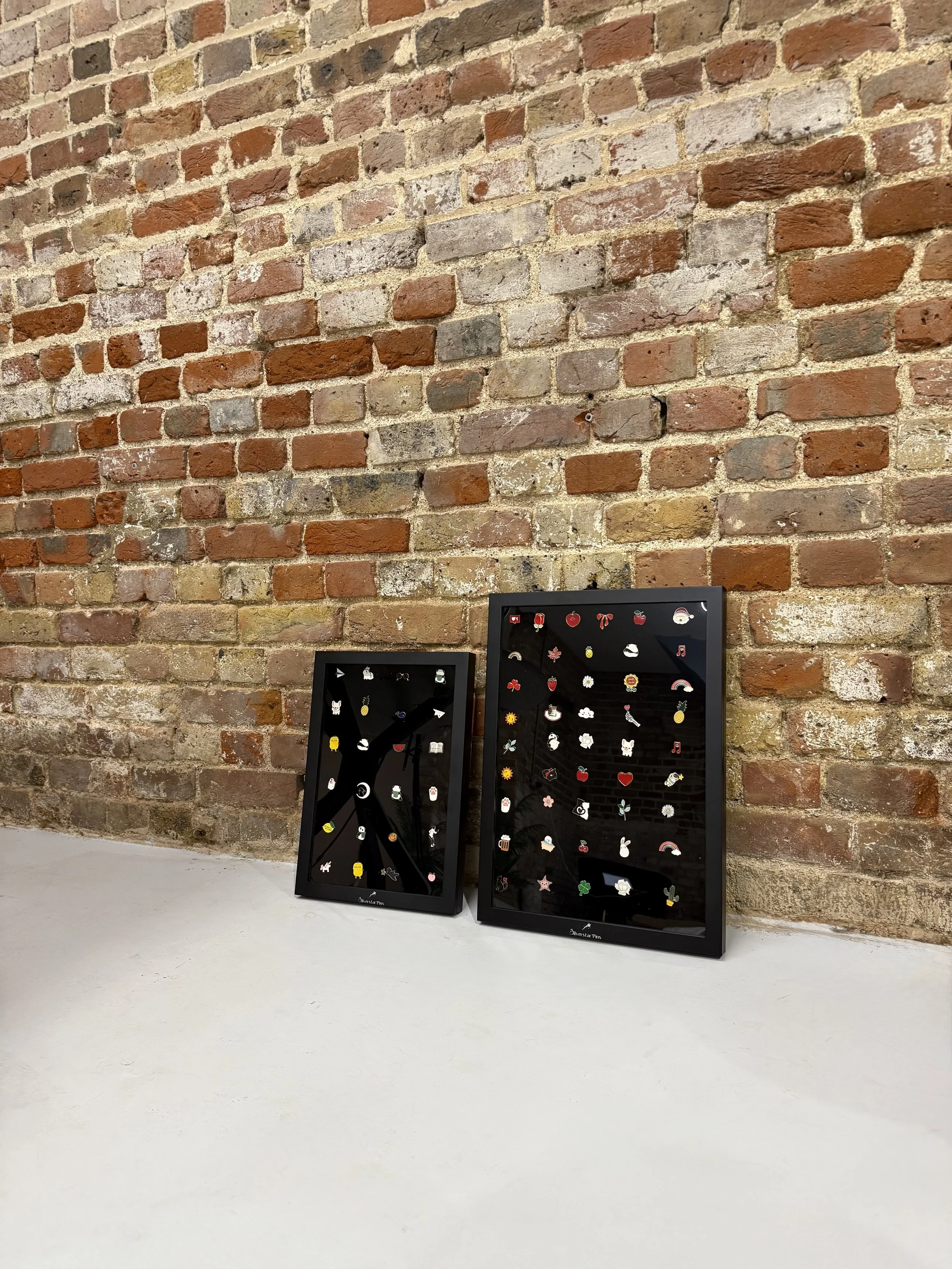 Two black display frames with small colorful pins or badges on a white surface, set against a brick wall background.
