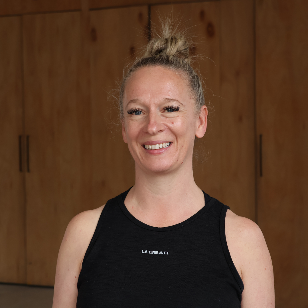 A smiling woman with blonde hair in a high bun, wearing a black sleeveless workout top, standing in front of wooden cabinets.