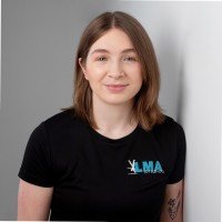 A young woman with shoulder-length brown hair wearing a black T-shirt with a logo, standing against a plain light background.