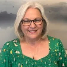 A woman with shoulder-length gray hair and glasses, smiling. She is wearing a green dress with yellow and white patterns, standing in front of a blurred outdoor background.