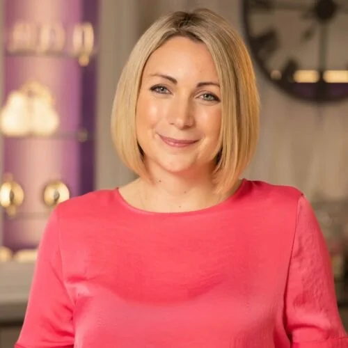 A smiling woman with shoulder-length blonde hair, wearing a pink top, indoors in a warmly lit room.