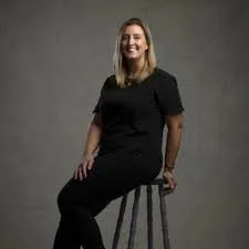 A woman with shoulder-length blonde hair sitting on a stool in front of a gray background, wearing a black dress and smiling.