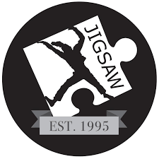 A black circular emblem featuring a person performing a gravity-defying stunt on a skateboard, with the words "JIGGSAM" and "EST. 1995".