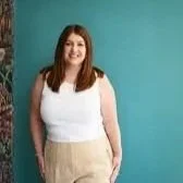 Woman with red hair wearing a white top and beige pants, smiling, standing against a teal background.