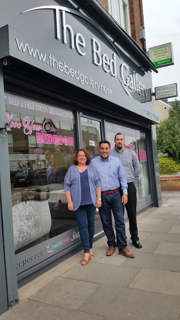 Three people standing in front of a store called The Bed Gallery, smiling for the photo. The store has large windows with pink lettering advertising free delivery and bed deals.
