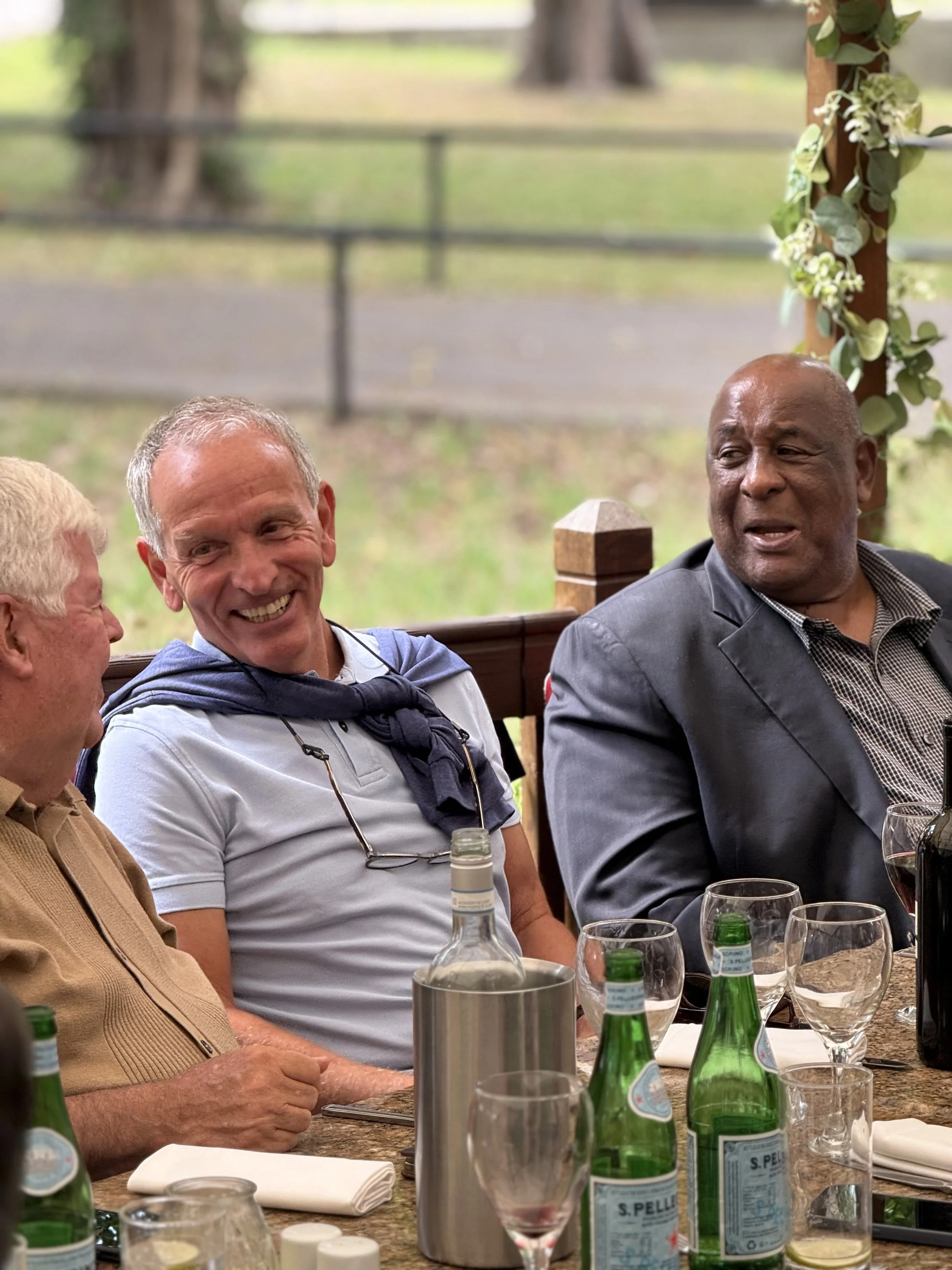 Three men are sitting at a table outdoors, engaging in conversation and smiling. The table has wine glasses, bottles of San Pellegrino sparkling water, and a metallic ice bucket. The background shows a green, park-like setting with trees and a fence.