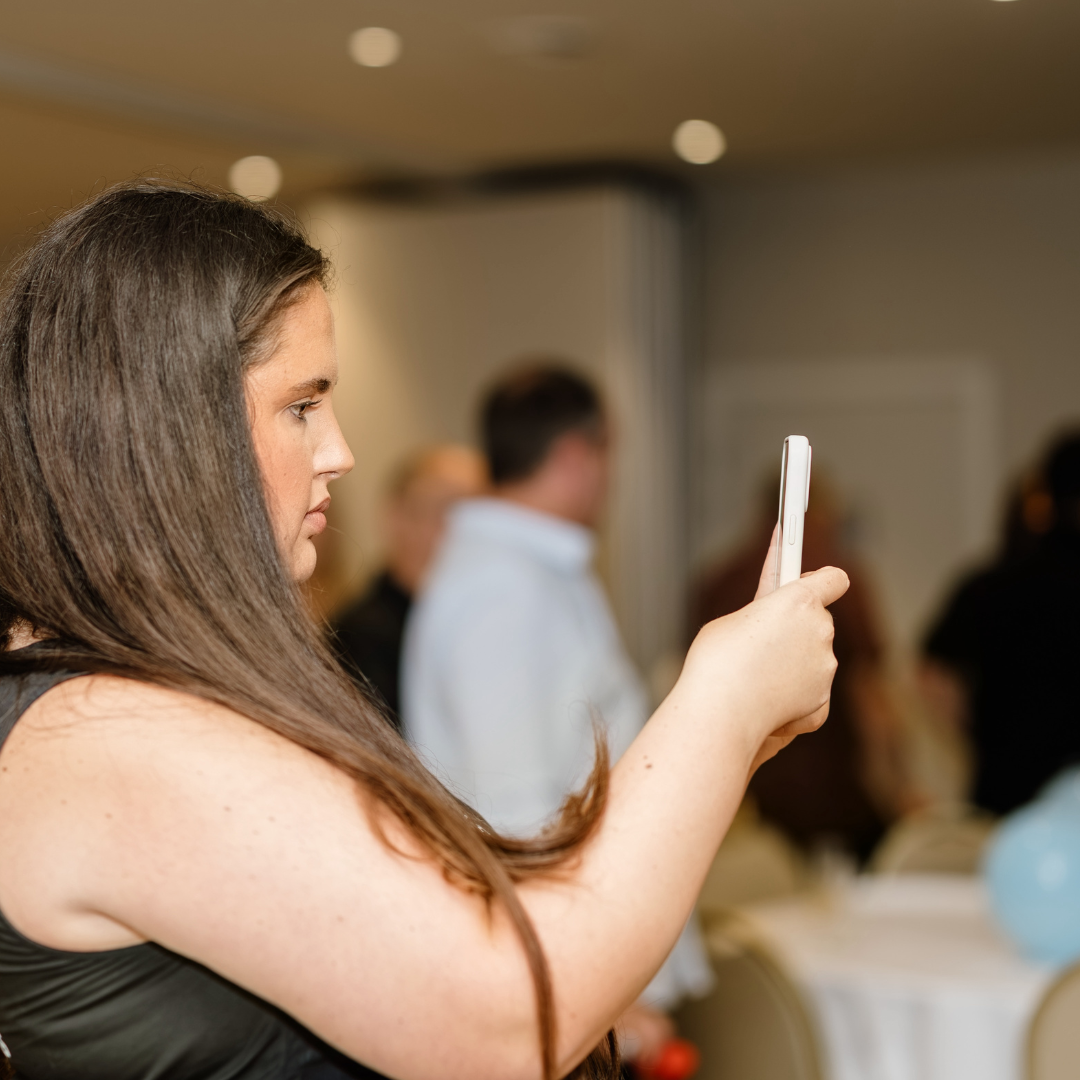 A woman with long brown hair taking a photo or video with her smartphone at an indoor event.
